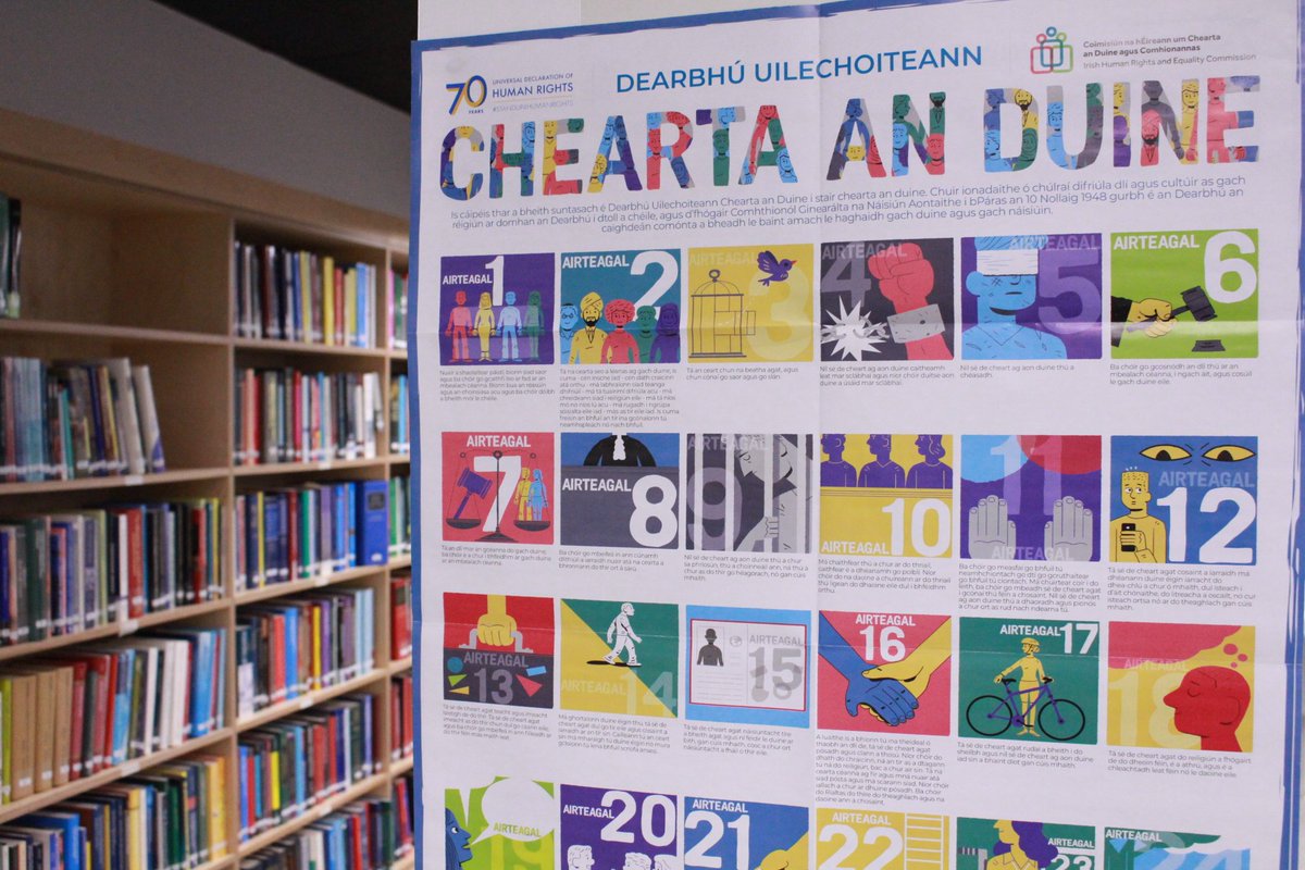 A poster hanging in a library. The colorful poster hangs portrait style and features separate illustrations for each of the 30 articles of the Universal Declaration on Human Rights.