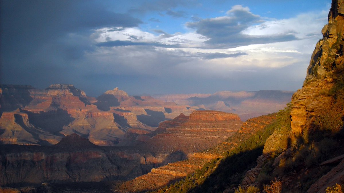 [Image description: colorful peaks and buttes take on orange tones from late afternoon light. Beyond a sheer cliff in the foreground, a vast desert landscape is revealed beneath a partially cloudy blue sky.