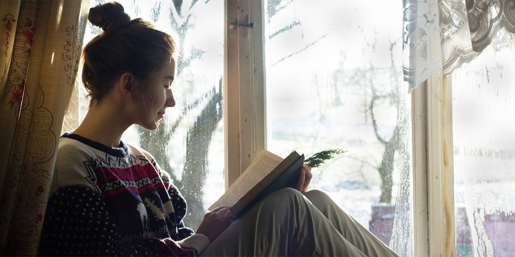 A young woman reads a book next to a rain-soaked window