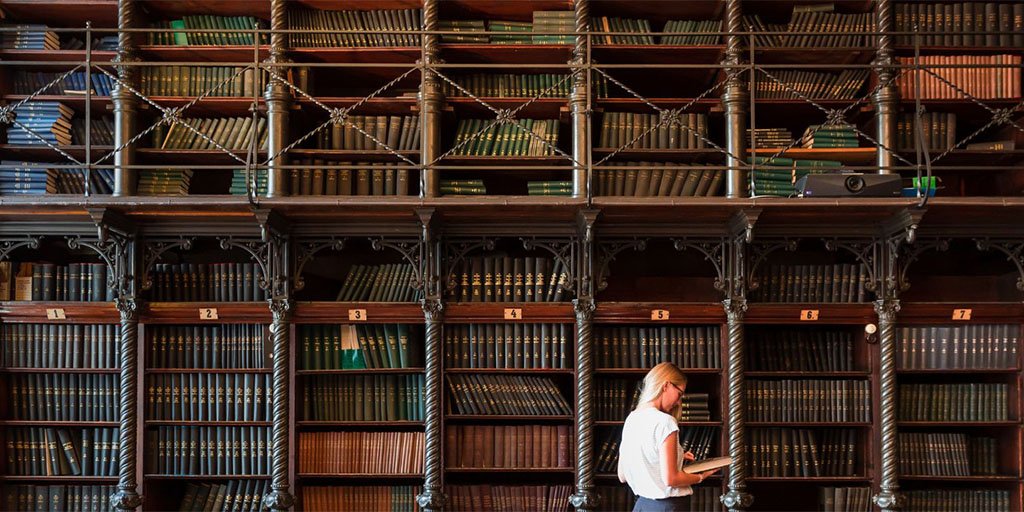 A woman stands in front of two stories of bookshelves in an old library