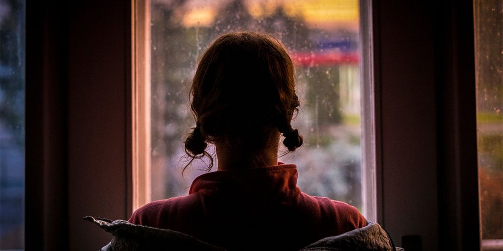A woman with knotted pigtails looks out a window, her jacket falling off her shoulders