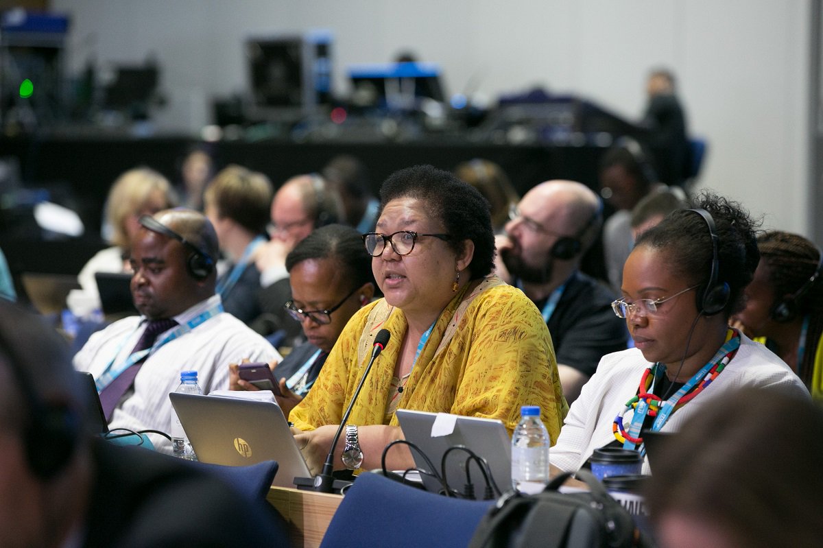 African woman delegate takes the mic at the ITU Plenipotentiary Conference surrounded by delegates, and technical equipment in the background