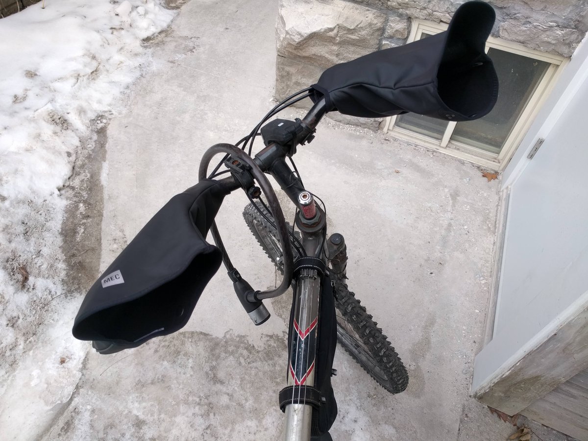 Photo looking downwards at bike handlebars with two "pogies" attached, neoprene mittens that attach to either end of the handlebar to keep your hands warm.