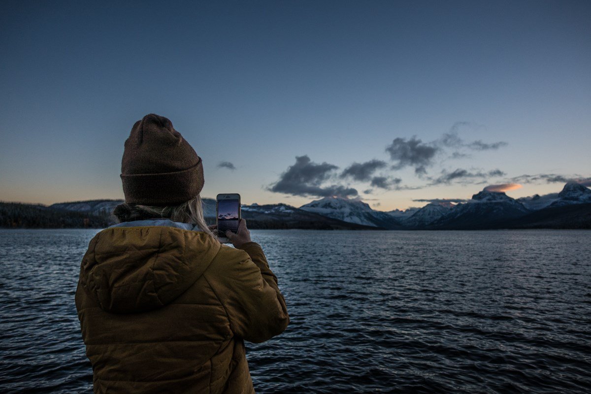 A person photographs sunrise over a lake with mountains in the background.