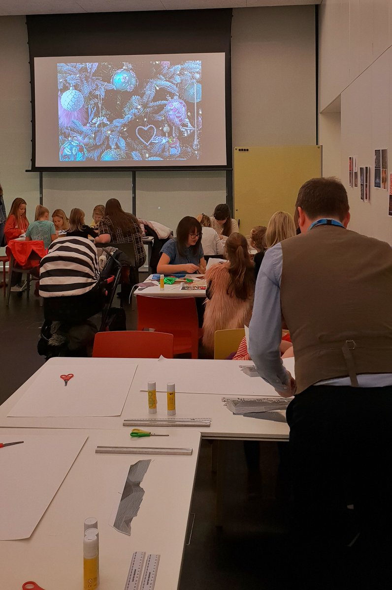 Workshop filled with people of different ages. Tables have card and glue on them. A screen above them shows Christmassy things like close-ups of a Christmas tree covered in decorations.