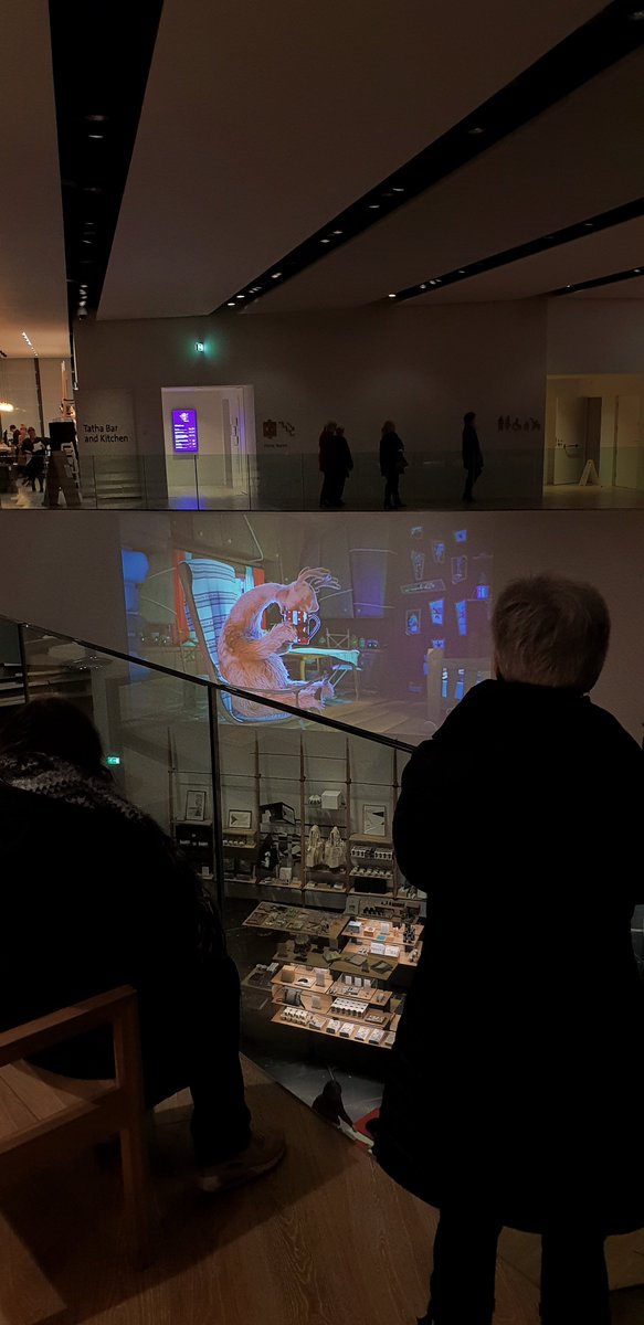 films projected on the wall above the shop in the main hall of V&A Dundee with people watching it from the balcony. The film on screen shows an animated moose in a chair burying its face in a mug.
