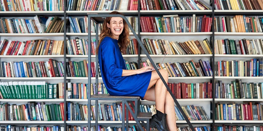A smiling woman sits on a library step ladder in front of stocked bookshelves