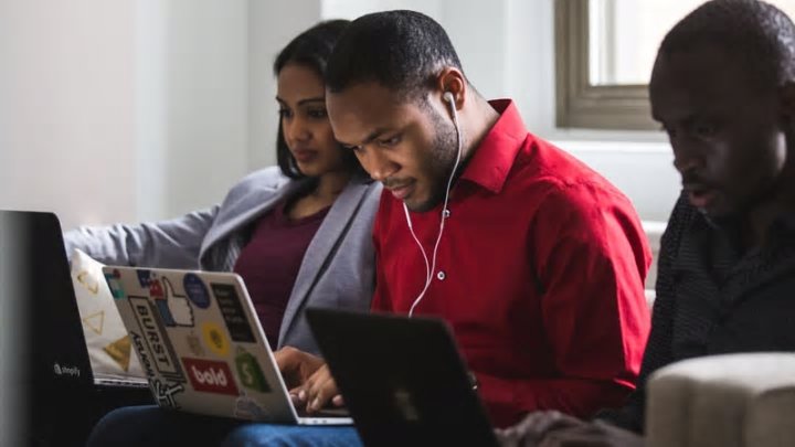 A man in a red shirt and wearing head-phones types on computer covered in stickers. He sits next to two other people on a bench in front of a light-filled window. 