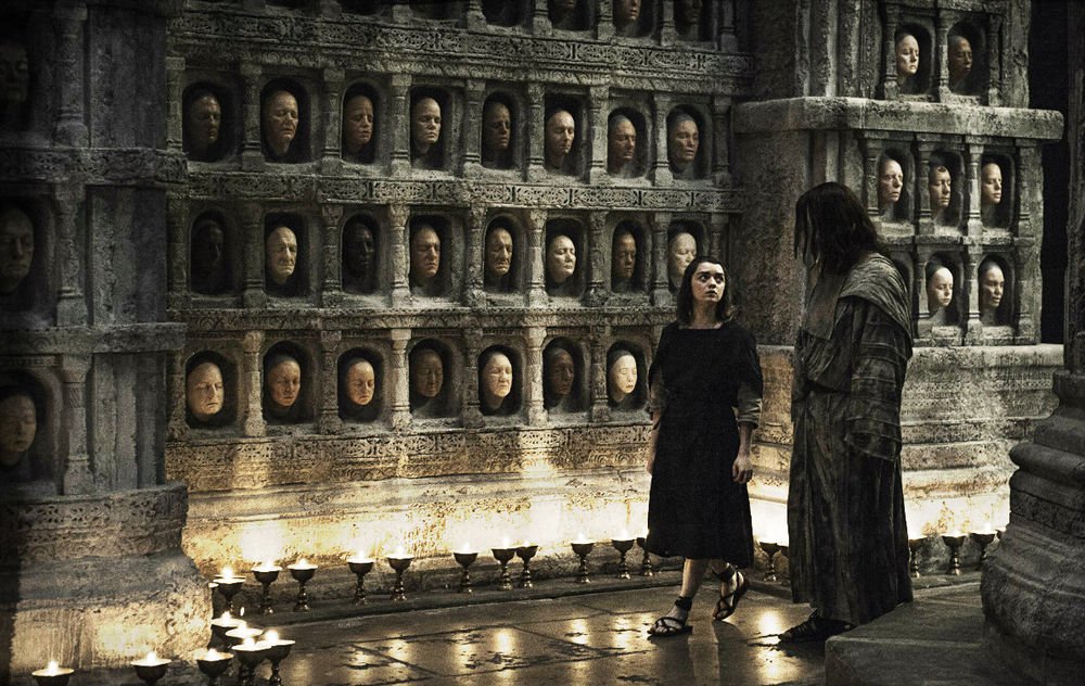 Arya Stark walking in a stone pillared room full of small alcoves. Each small alcove has a human face skinned from someone.