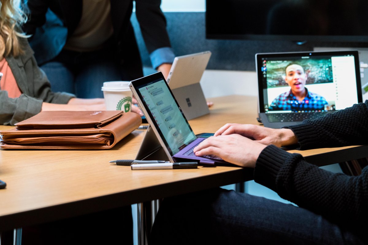 An employee is typing on a Microsoft Surface with a purple keyboard. There is another Surface open next to him showing a video call. There are two other employees leaning over the table on the other side working on a third Surface, but their faces are not pictured. 