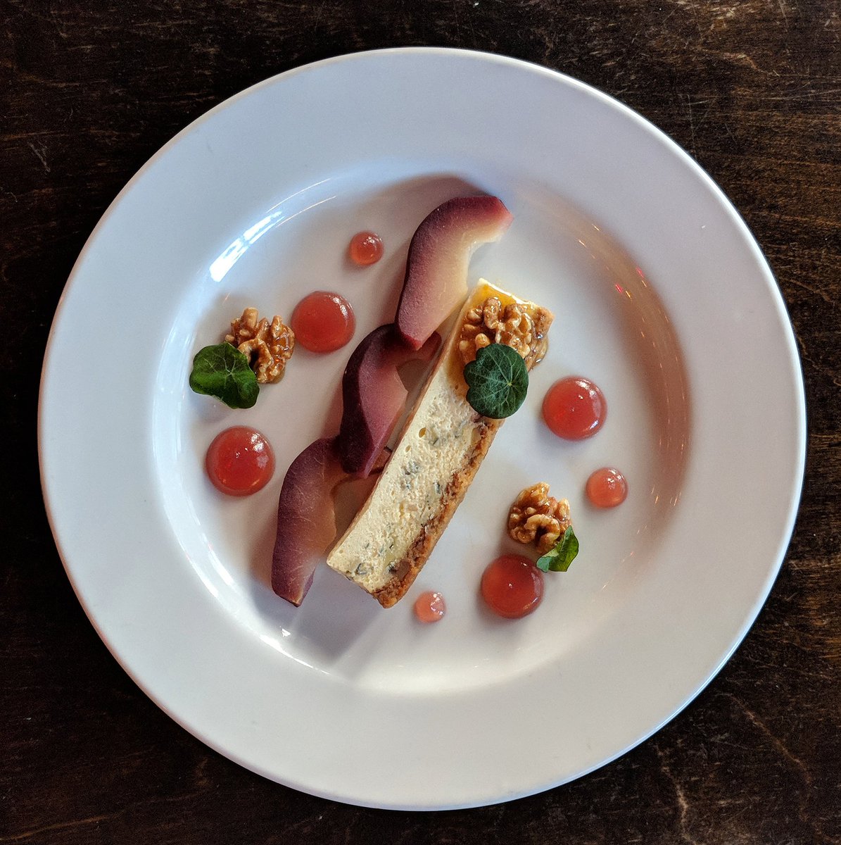 Bleu d'Auvergne Cheesecake (Hint: it's actually intended as a cheese course, not a traditional dessert. 🧀) It's made with Bleu d'Auvergne cheese, house-made graham cracker crust, honeyed walnuts, red wine poached pears, red wine coulis, and nasturtium leaves. 🌿🍰🧀