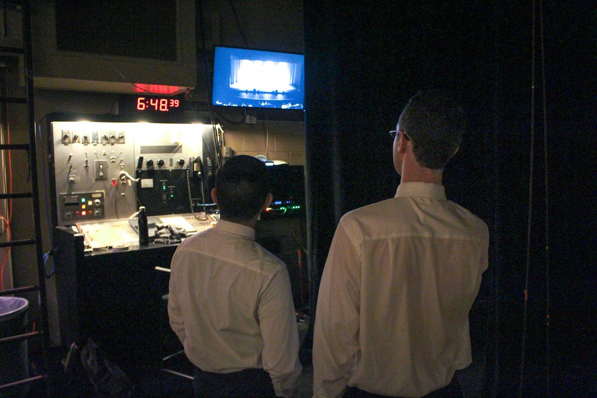 Photo from the backstage of a theater. The sources of light are on the background wall: bright bulbs above a stage manager's panel, a digital clock reading "6:48.39" in red LED, and a flat screen TV showing a camera feed of the stage. Two young adults stand in the foreground, visible from the waist-up. Dressed in white long-sleeve button-downs and black pants, they face back at the clock and TV.