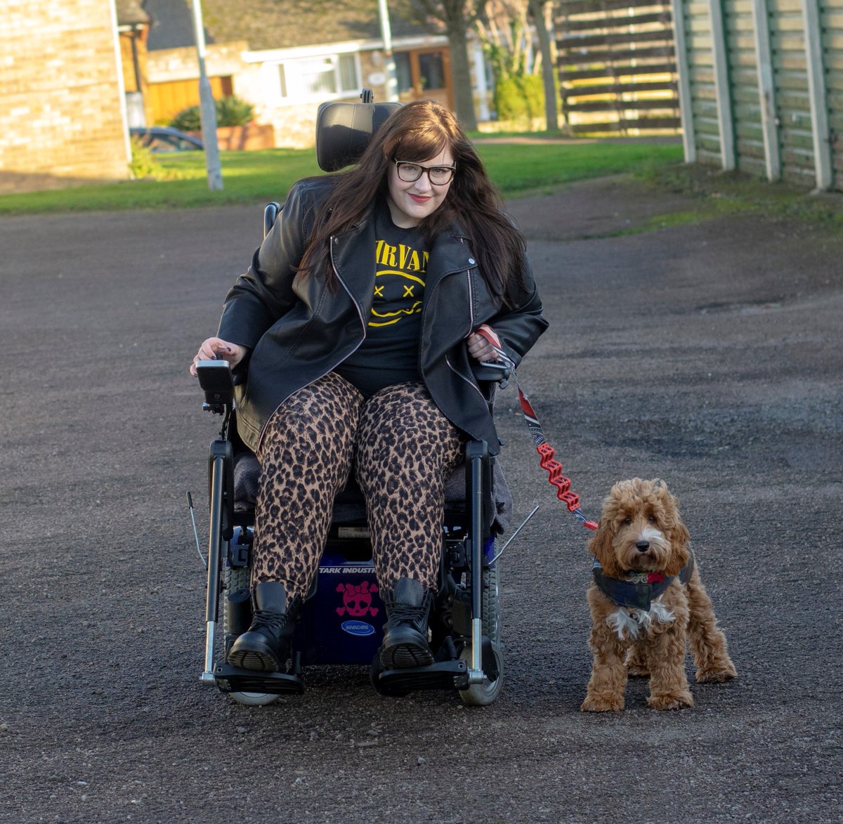 Me sat in my powerchair wearing Nirvana tshirt, leather jacket and leopard print trousers holding lead of red and white cockapoo puppy