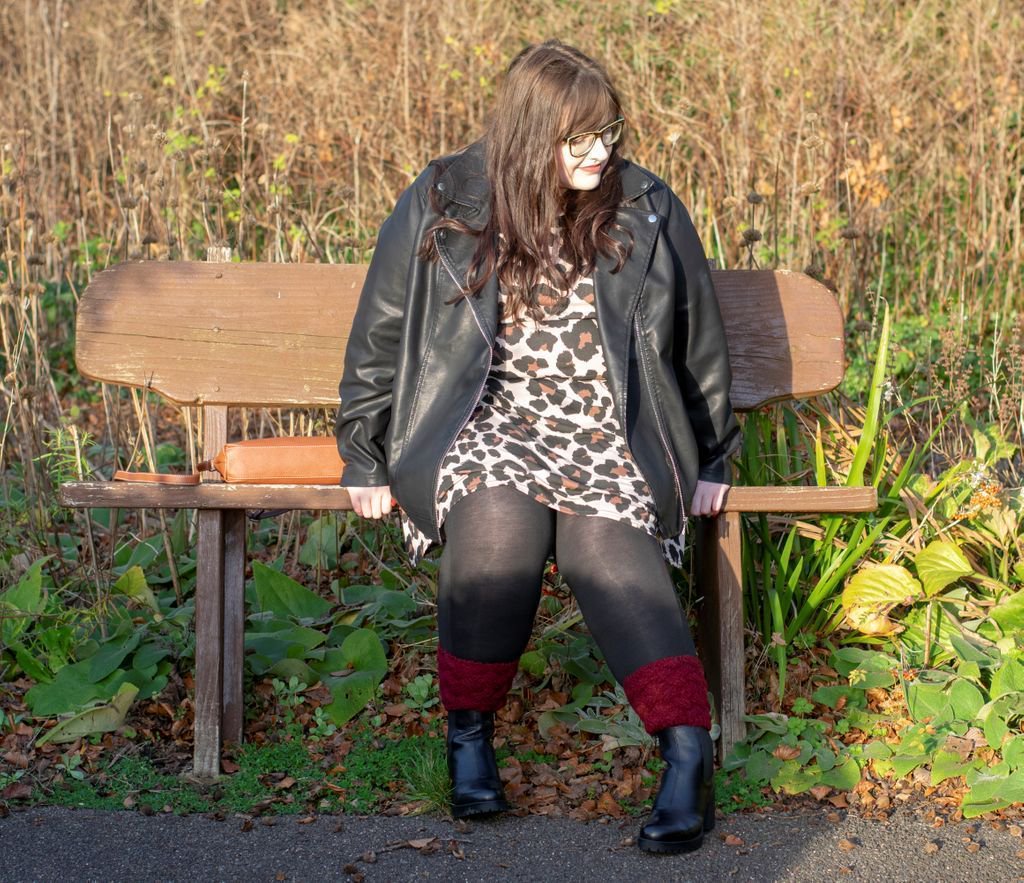 Woman sat on bench with long brown hair down, wearing leopard print dress, leather jacket, black tights and black Chelsea boots with red ankle cuffs. Greenery in background
