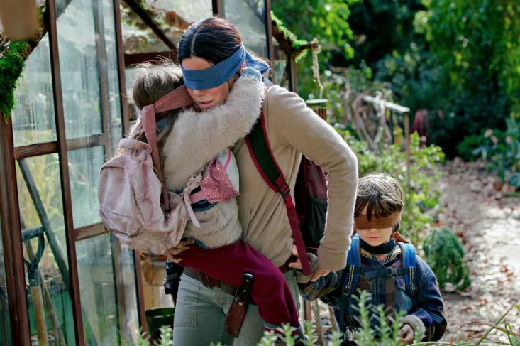 Sandra Bullock wearing a blindfold carrying a child who is blindfolded and holding the hand of another child who is blindfolded as they walk 