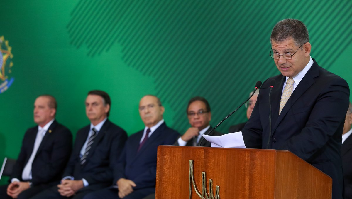 #PraCegoVer: Foto mostra o ministro da Secretaria-Geral da Presidência, Gustavo Bebianno, discursando ao microfone em cerimônia no Palácio do Planalto. Ao fundo estão sentados, da esquerda para a direita, o ministro da Casa Civil, Onyx Lorenzoni, o presidente da República, Jair Bolsonaro, o ex-ministro da Casa Civil Eliseu Padilha e o ex-ministro da Secretaria-Geral da Presidência Ronaldo Fonseca.