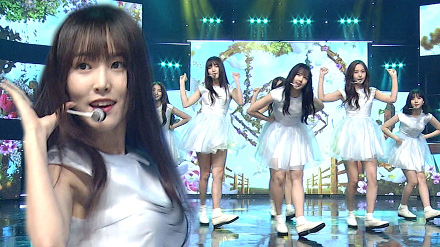 A close-up of a young woman with long dark hair wearing a headset and white outfit on the left, with a group of similarly dressed women performing a synchronized dance in white dresses on a brightly lit stage with floral decorations in the background.