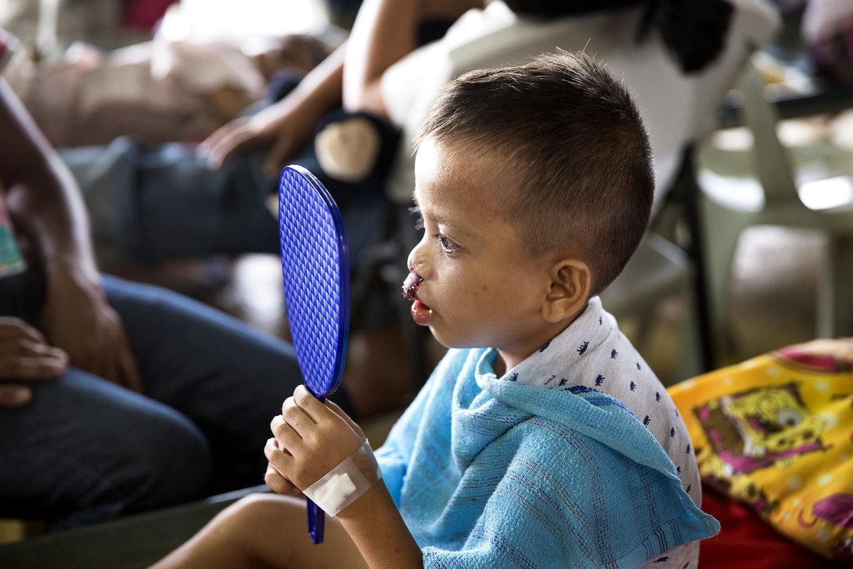 A small child sitting on a table is holding a mirror looking at his face. A blue towel is draped over the front of him and in the background are other people sitting on chairs. 