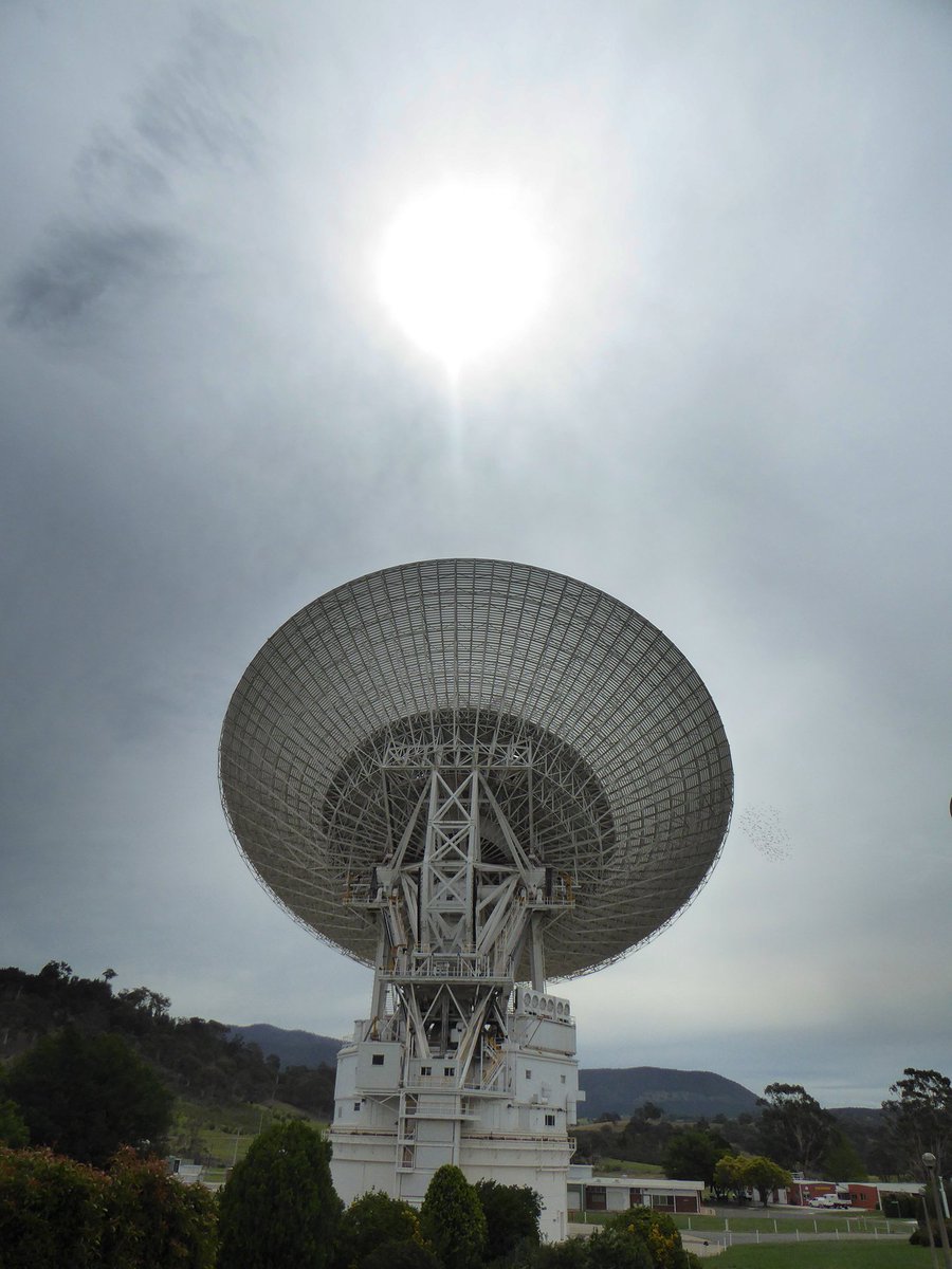 Sun shining through clouds above deep space antenna dish.