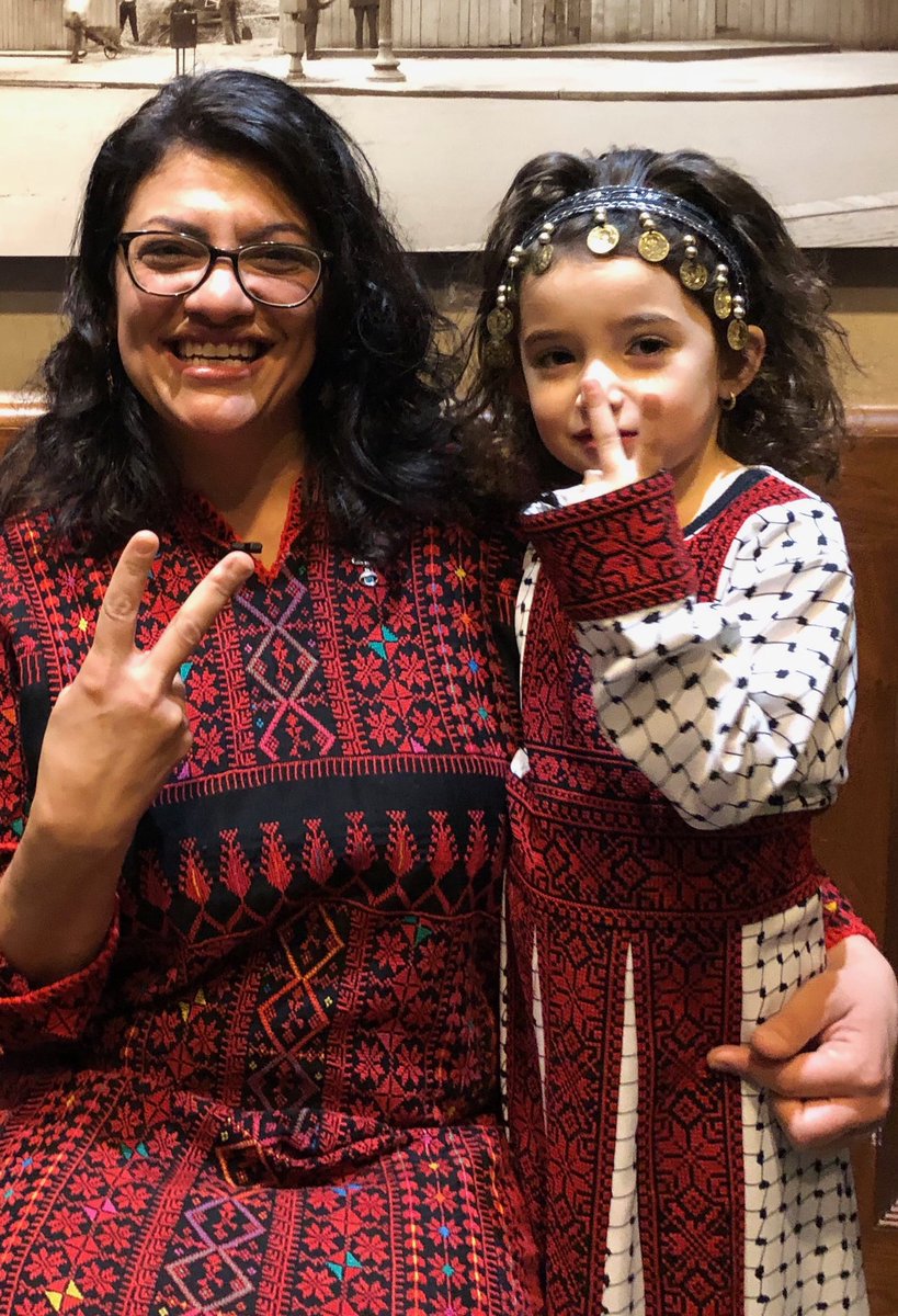 New Palestinian-American Congresswoman Rashida Tlaib, from Detroit Michigan, wearing her traditional embroidered dress (thobe) & holding her young daughter dressed in traditional attire. Both are holding up peace signs with their hands and looking very happy.