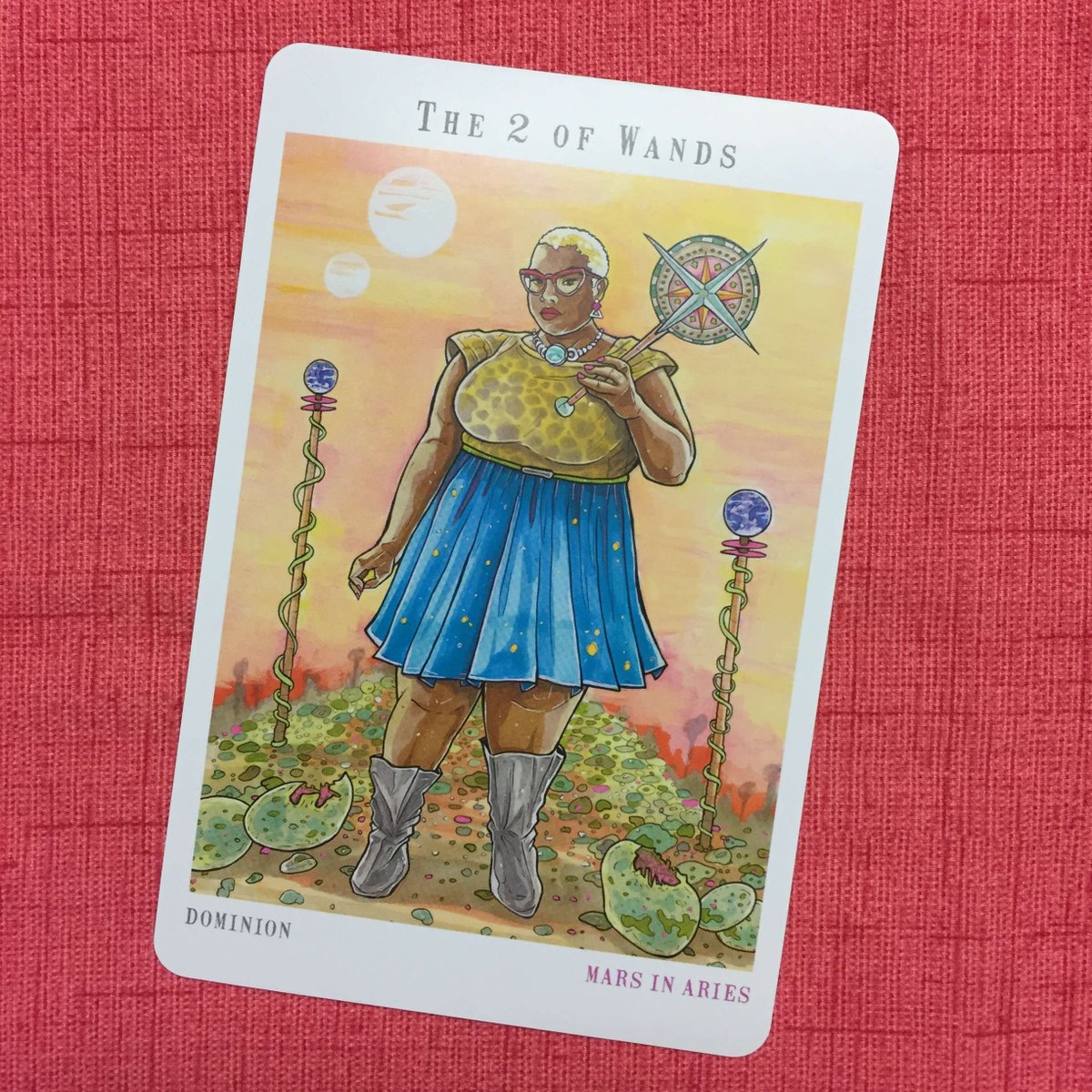 Next World Tarot - Two of Wands - A Black femme in a blue skirt and a leopard print top, with close-cut hair, holds a mace in her left hand and regards the reader through cat-eye glasses.