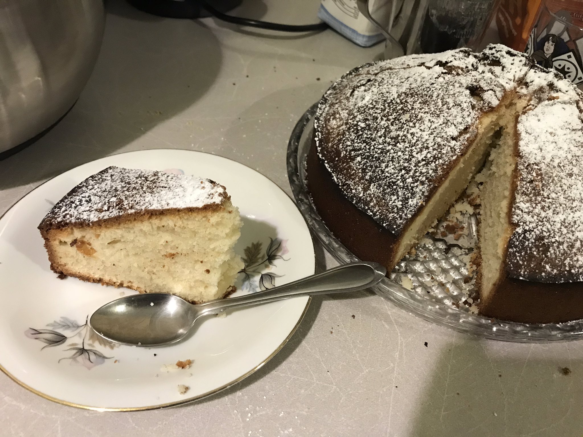 A round cake, looks like a sponge cake with powdered sugar on top, with a section cut out and put on a plate beside it. The cake has a rather thick, dark brown crust.