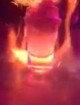 Screenshot from a video, with colors consisting entirely of fiery hues (between red and yellow). A filled shot glass sits on a table as flames dance around it. Empty space underneath the glass bottom allows one to clearly see the flames behind it.