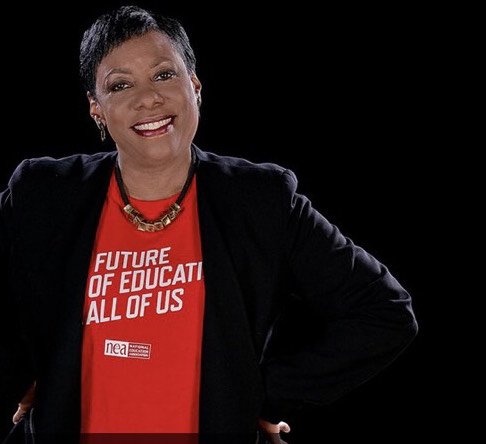 Becky Pringle, Vice President of the National Education Association smiling in front of a black background wearing a black blazer and a red shirt which reads "THE FUTURE OF EDUCATION IS ALL OF US."