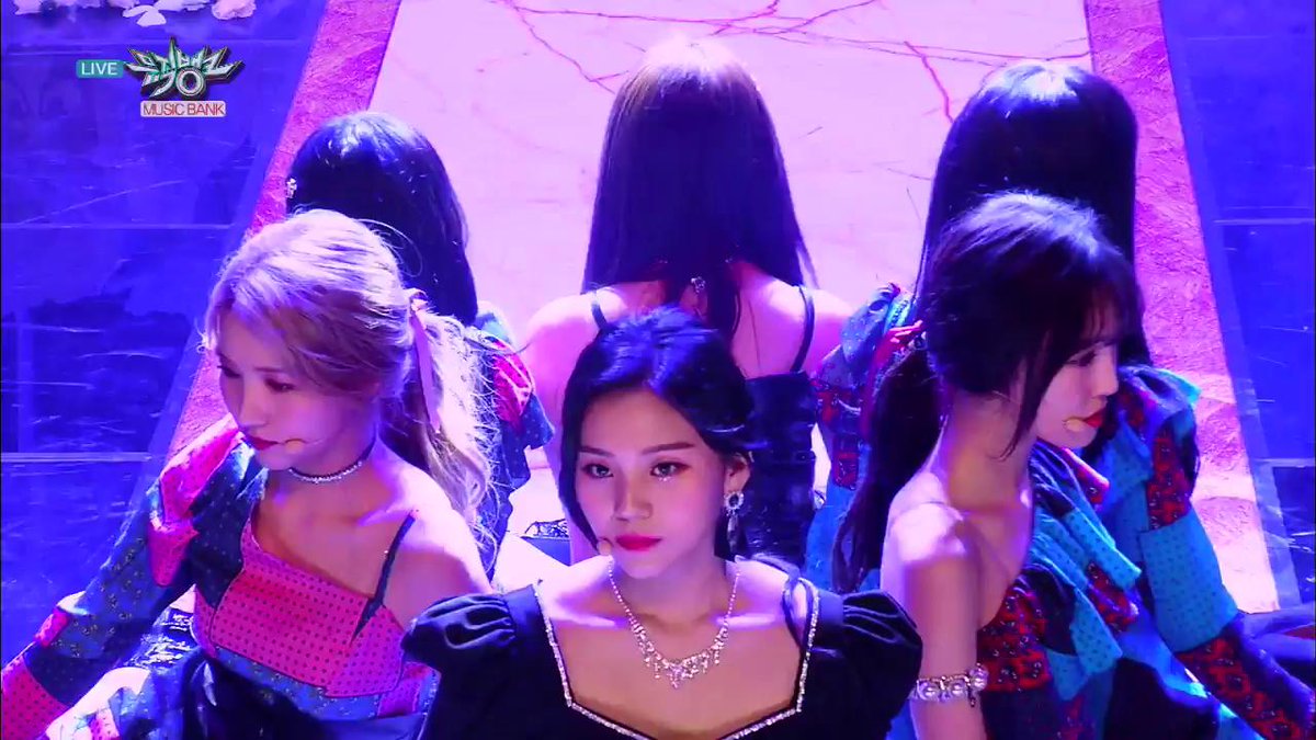 Screenshot from a KBS2 "Music Bank" live broadcast showing K-pop female performers in pink and blue stage outfits on a stage with purple lighting.