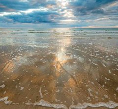 Small ocean waves wash up on a wide sandy beach under a cloudy morning sky.