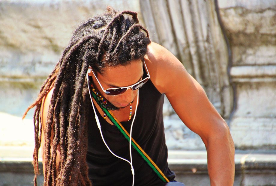 African man wearing long dreads, sunglasses, tank top, head down listening to smartphone on earbuds