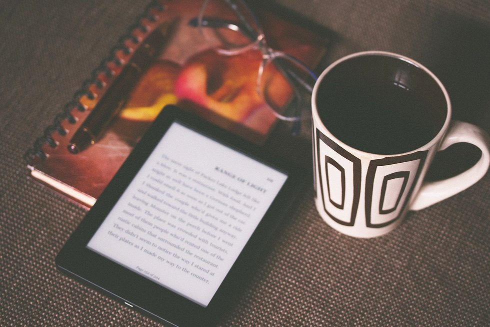 an eBook reader on a table beside a notebook and a cup of coffee