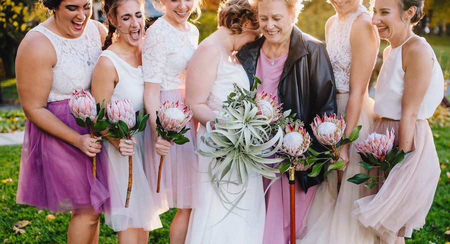 This bride's bridesmaids—including her grandmother (and maid of honor), Jan—carried king protea flowers that matched their purple and blush tulle skirts from Revelry. See more from this adorable celebration here: bit.ly/2UNlxN9