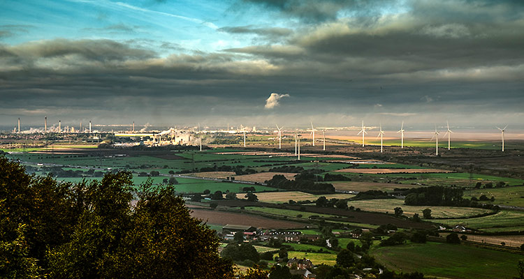 The Ince Marshes site. In the distance you can see a wind farm and above that to the right grey clouds are rolling in. On the left the sky is still blue. In the foreground are loads of fields in beige and green and you can also see hedgerows. 