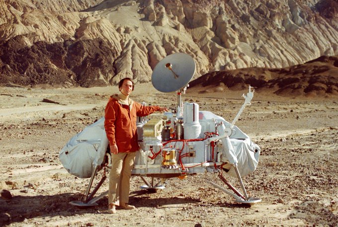 Carl Sagan standing next to a Viking spacecraft in a rocky and desolate environment.