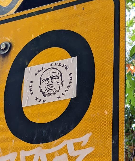 White sticker on a yellow road sign that says "All Cops Are Derek Chauvin"
And has Chauvins face in the middle