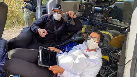 "Queenpins" directors Aron Gaudet, left, and Gita Pullapilly on set. both are wearing masks while watching footage on a screen.