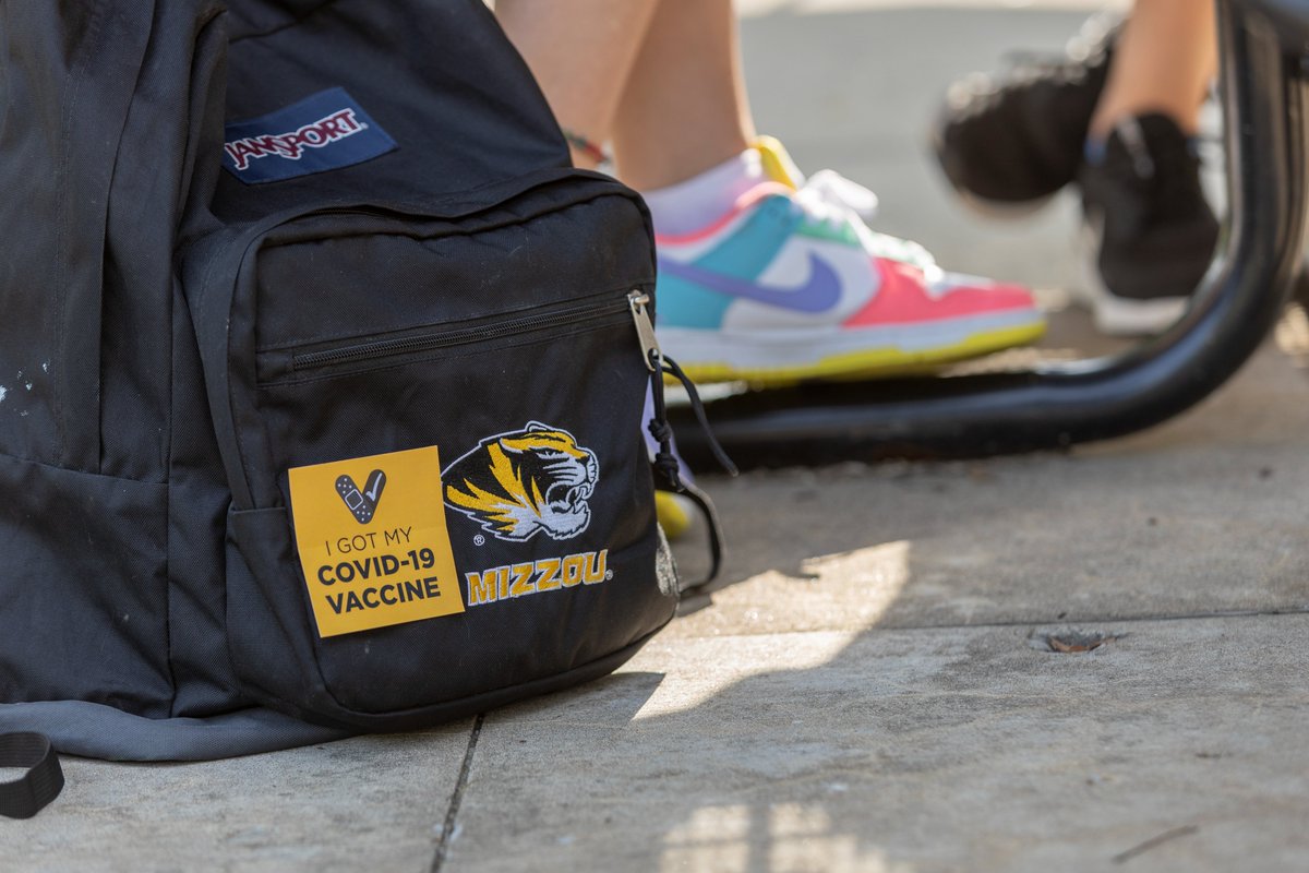 A backpack under an outdoor table is photographed. The legs and shoes of two students are visible in the background. 