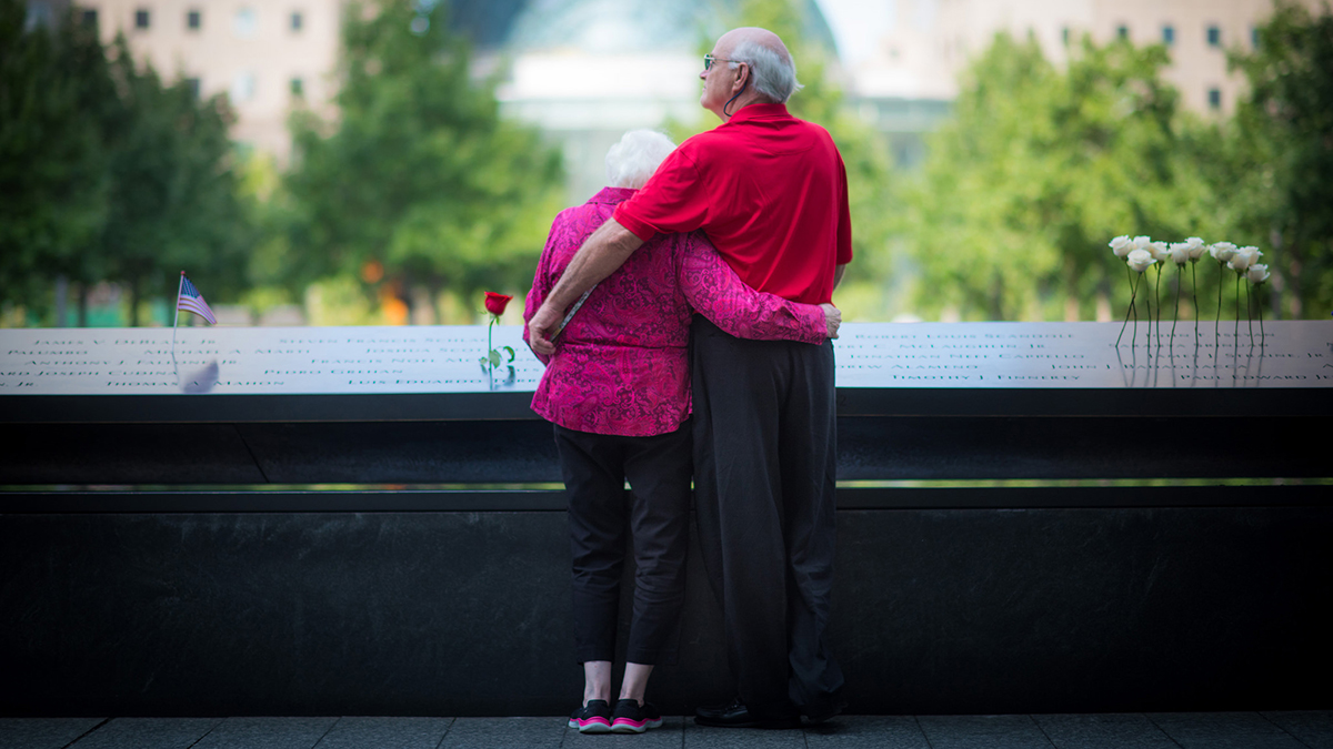 An elderly White couple stand with their arms around each other while paying their respects at the September 11th memorial.