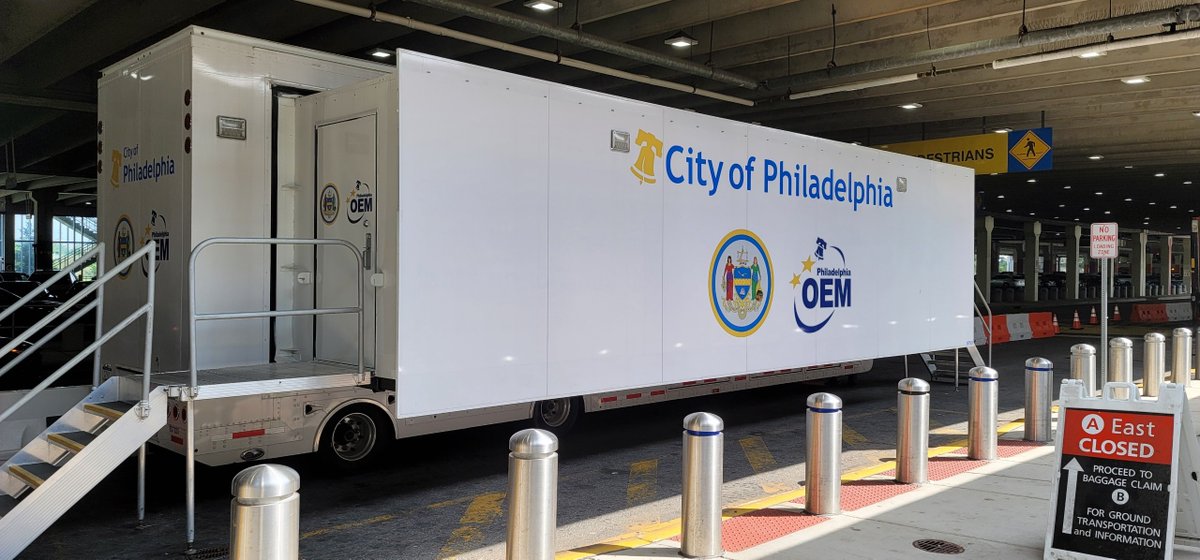 large trailer labeled "city of philadelphia OEM" parked outdoors near bollards under concourse at airport