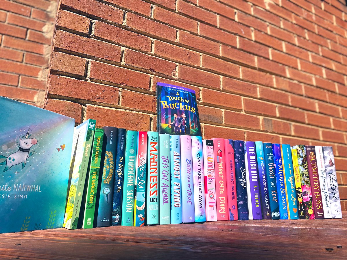 A rainbow of book spines on a red brick backdrop. The books shown are Not Quite Narwhal by Jessie Sima, Taproot by Kerry Young, The Magic Fish by Trung Le Nguyen, Strangeworlds Travel Agency by L.D. Lapinski, Too Bright to See by Kyle Lukoff, Hurricane Season by Nicole Melleby, Girl from the Sea by Molly Ostertag, That Way Madness Lies edited by Dahlia Adler, Jay’s Gay Agenda by Jason June, Almost Flying by Jake Arlow, Both Can Be True by Jules Machias, Can’t Take That Away by Steven Salvatore, The Unpopular Vote by Jasper Sanchez, Meet Cute Diary by Emery Lee, Between Perfect & Real by Ray Stoeve, Starfish by Akemi Dawn Bowman, The Witch King by H.E. Edgmon, How to Become a Planet by Nicole Melleby, The Ghosts We Keep by Mason Deaver, The Mirror Season by AMM, All You Knead is Love by Tanya Guerrero, Snapdragon by Kat Leyh, Wild Beauty by AMM, Cemetery Boys by Aiden Thomas, The Passing Playbook by Isaac Fitzsimons, Elatsoe by Darcie Little Badger, A Touch of Ruckus by Ash Van Otterloo