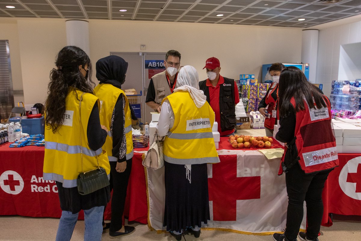 three women wearing vests labeled "interpreter" stand indoors in front of red cross tables with fruit and near four red cross staffers
