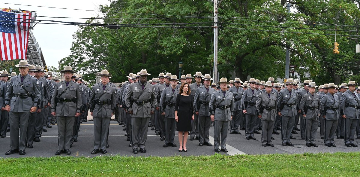 Governor Hochul stands outdoors wearing a black dress in line with New York State Police Troopers in uniform