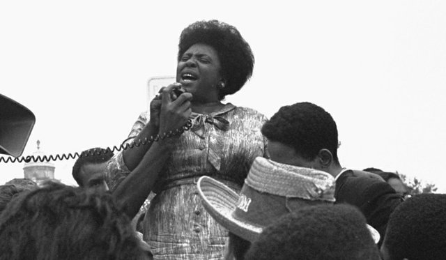 A black and white photo of civil rights activist Fannie Lou Hamer standing above a crowd of people and speaking into a microphone.