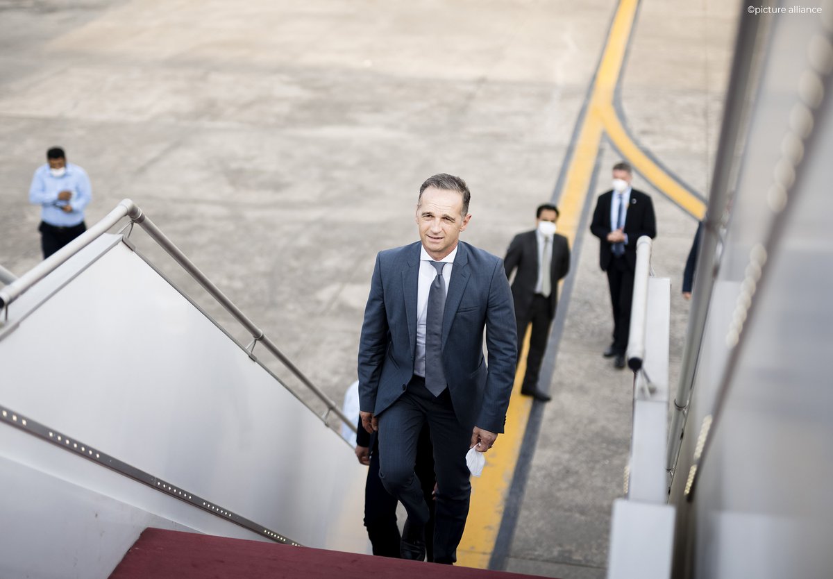 Minister Maas photographed as he ascends the maroon steps of a plane. A few men in suits and masks watch from below. 