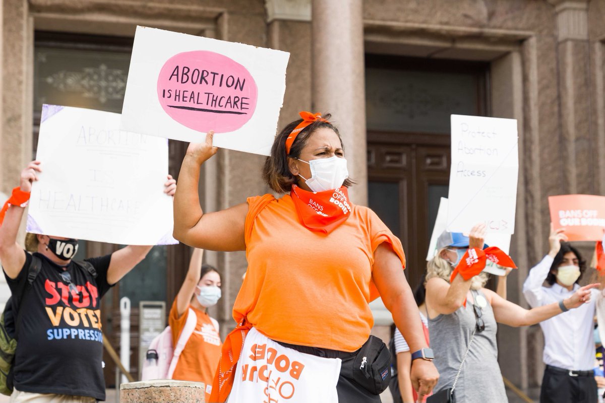 Person in an orange shirt wearing a mask standing at a protest in Texas. The person is holding a sign that reads "Abortion is healthcare"