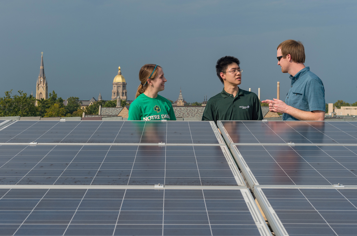 Three people tour a solar panel field on the roof of Stinson-Remick Hall at the University of Notre Dame. The Golden Dome and spire of the Basilica of the Sacred Heart can be seen behind them.