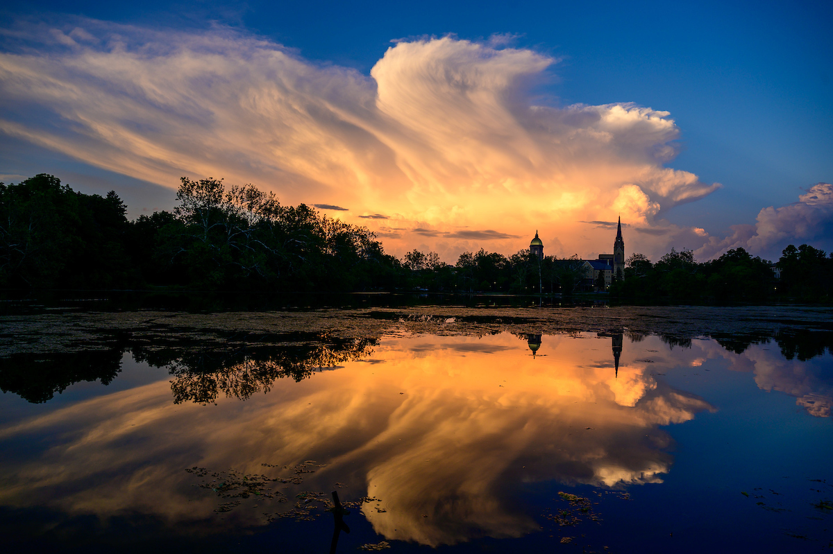 Distant storm clouds are lit golden by the sun at sunrise over St. Mary's Lake at the University of Notre Dame. The clouds and the Golden Dome and spire of the Basilica of the Sacred Heart are all reflected in the water.
