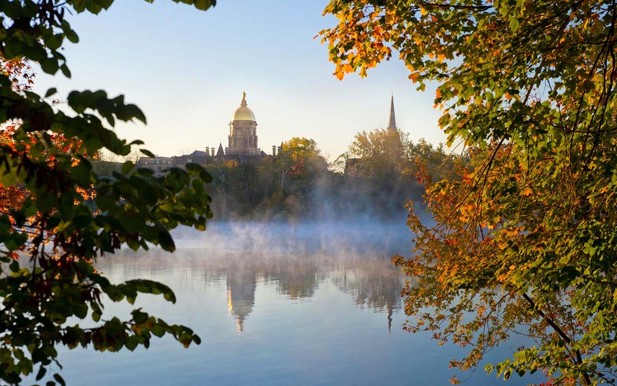 Mist rises on St. Joseph's Lake at the University of Notre Dame with the Golden Dome of the Main Building and the spire of the Basilica of the Sacred Heart on the other side. Both buildings are reflected in the still water, and green and orange fall foliage frames the scene.