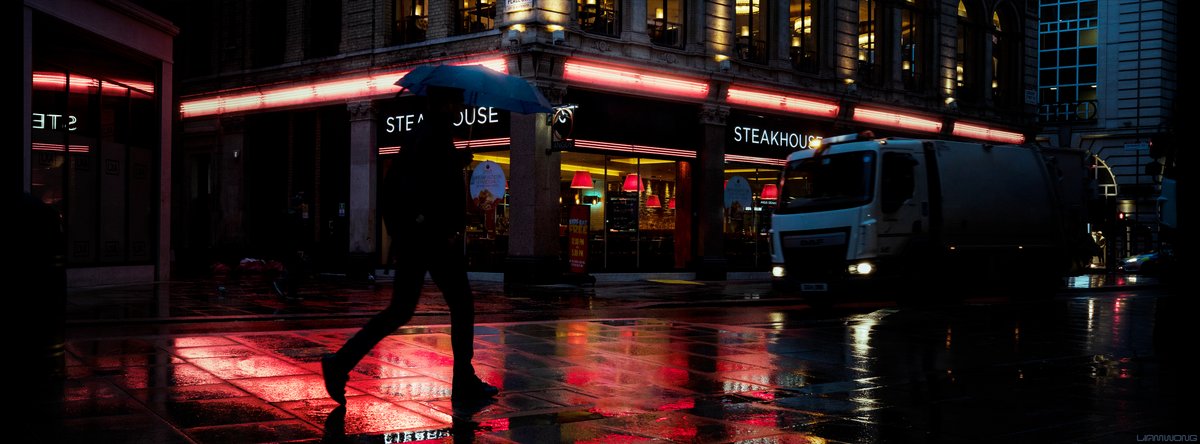 Photography by Liam Wong of London at night. A man walking through the rain, backlit by a red neon sign from a steakhouse. The pavement is lit up in red.